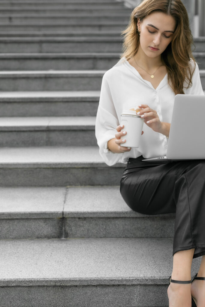 A business woman sitting on the steps with a cup of coffee and a laptop, comparing Flodesk vs. Mailchimp for her email marketing campaign.