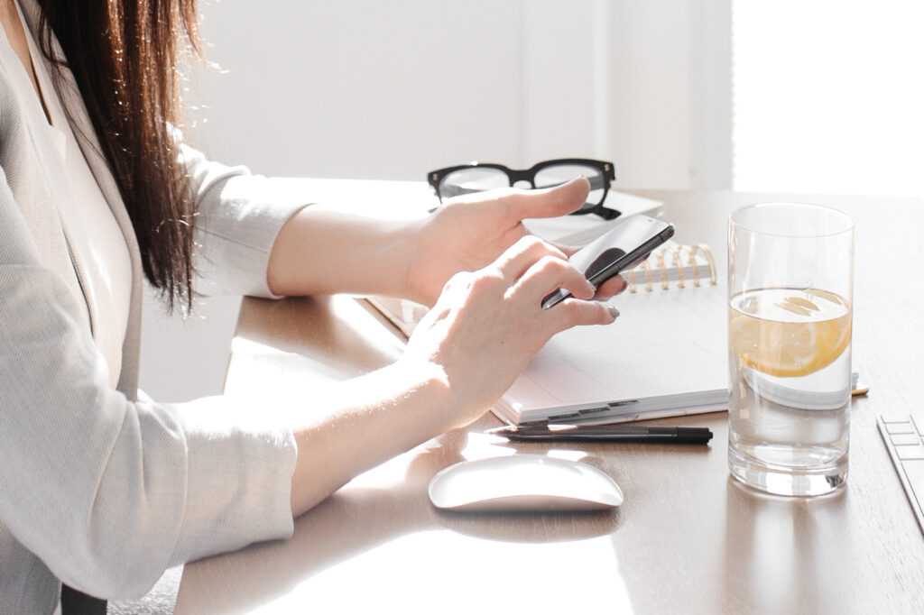 A woman sitting at a desk with a cell phone and a glass of water, comparing Honeybook vs. Quickbooks.
