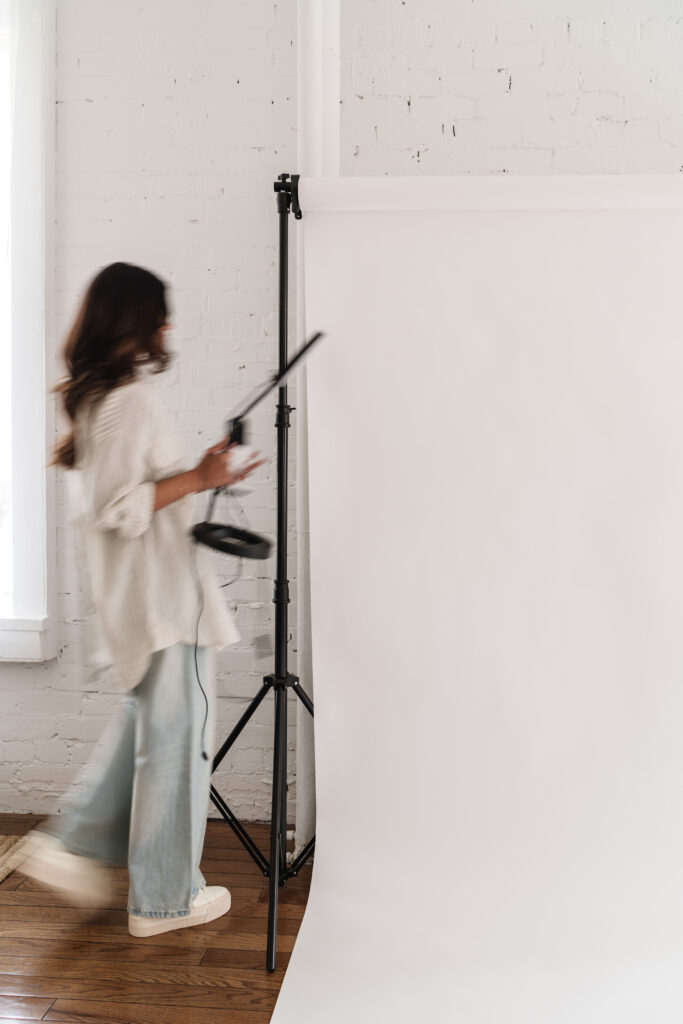 A woman holding a camera in front of a white backdrop, capturing images for social media.