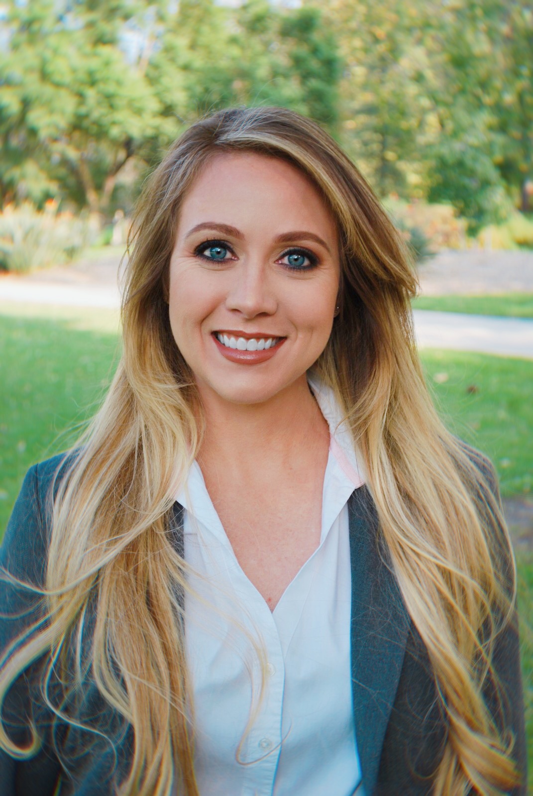 A woman with long blonde hair and blue eyes is smiling, standing outside in a park, wearing a white shirt and a grey blazer, perhaps on her way to a networking event.