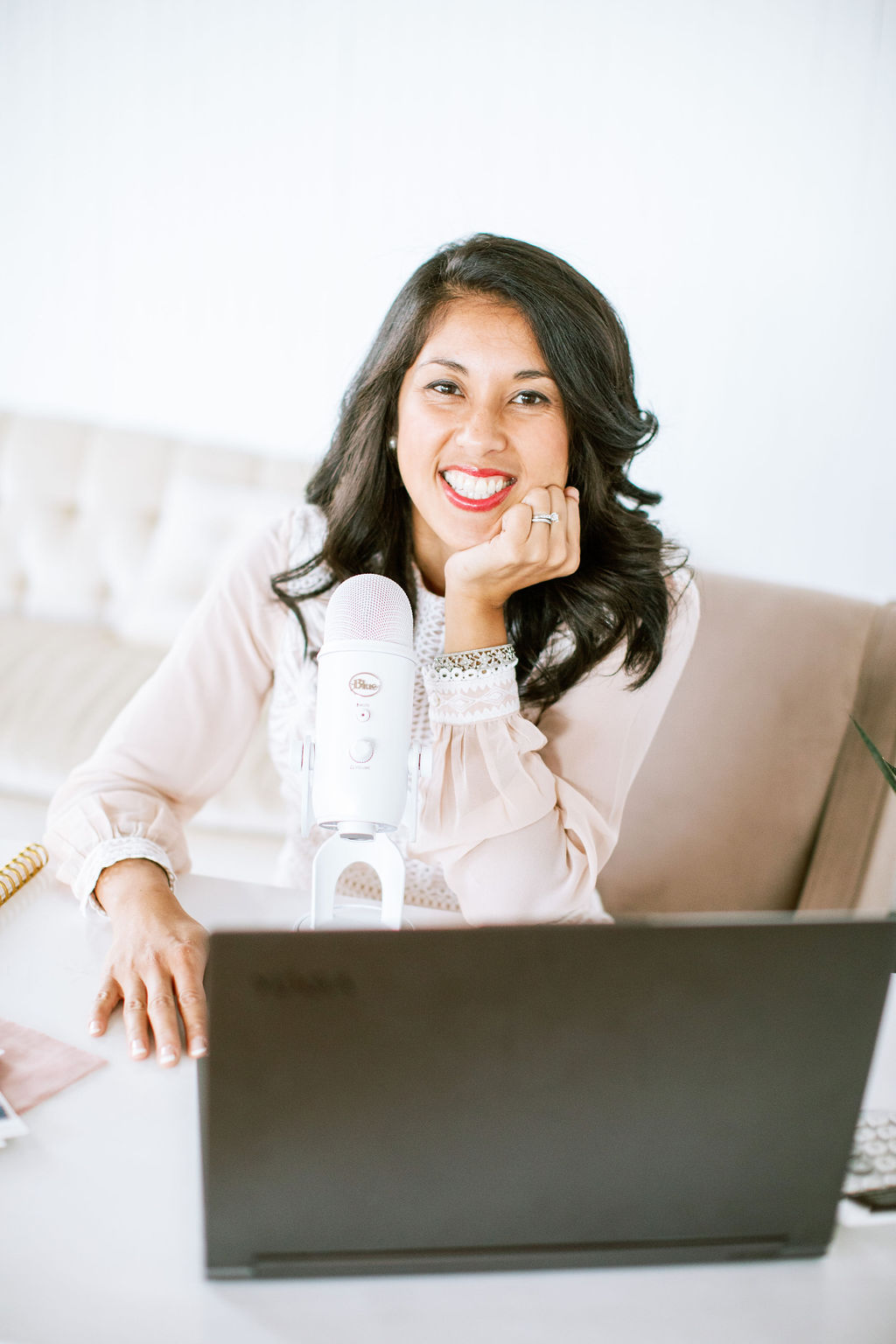 A woman with dark hair, smiling, sits at a desk with a laptop and a white microphone in front of her, starting a podcast.