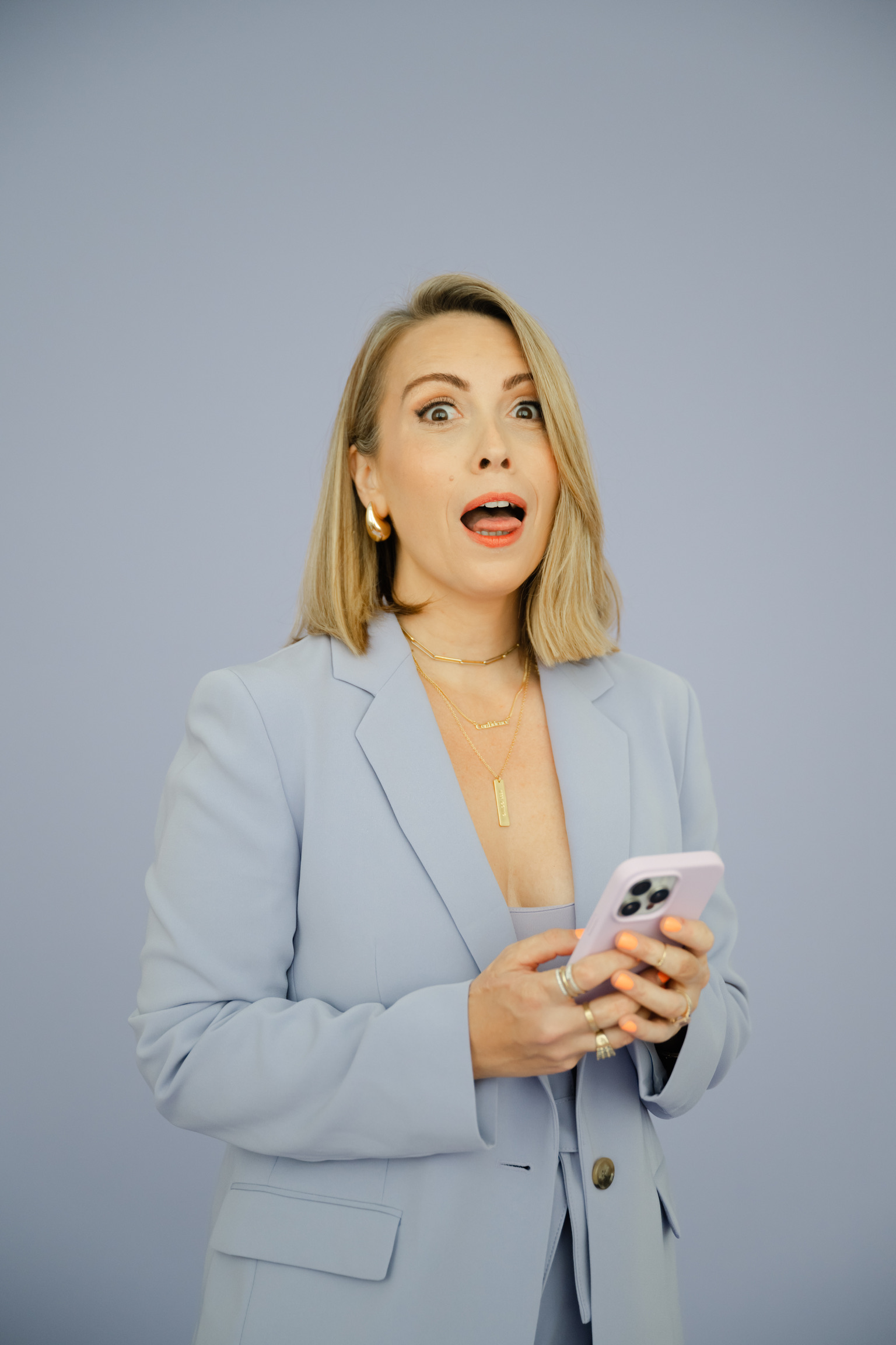 A woman with blonde hair in a light blue blazer holds a smartphone and looks surprised against a plain blue background, perhaps struck by an innovative Instagram strategy for wedding pros.