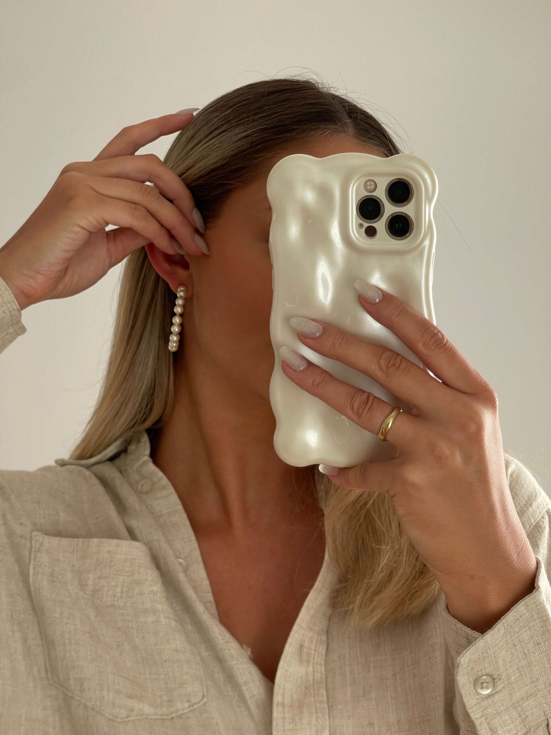 A person with long hair, possibly a creative wedding planner, holds a phone with a unique, bulbous case covering their face. Wearing a beige shirt, hoop earrings, and a ring on their left hand while adjusting their hair, they might be looking for canva templates to inspire their next project.