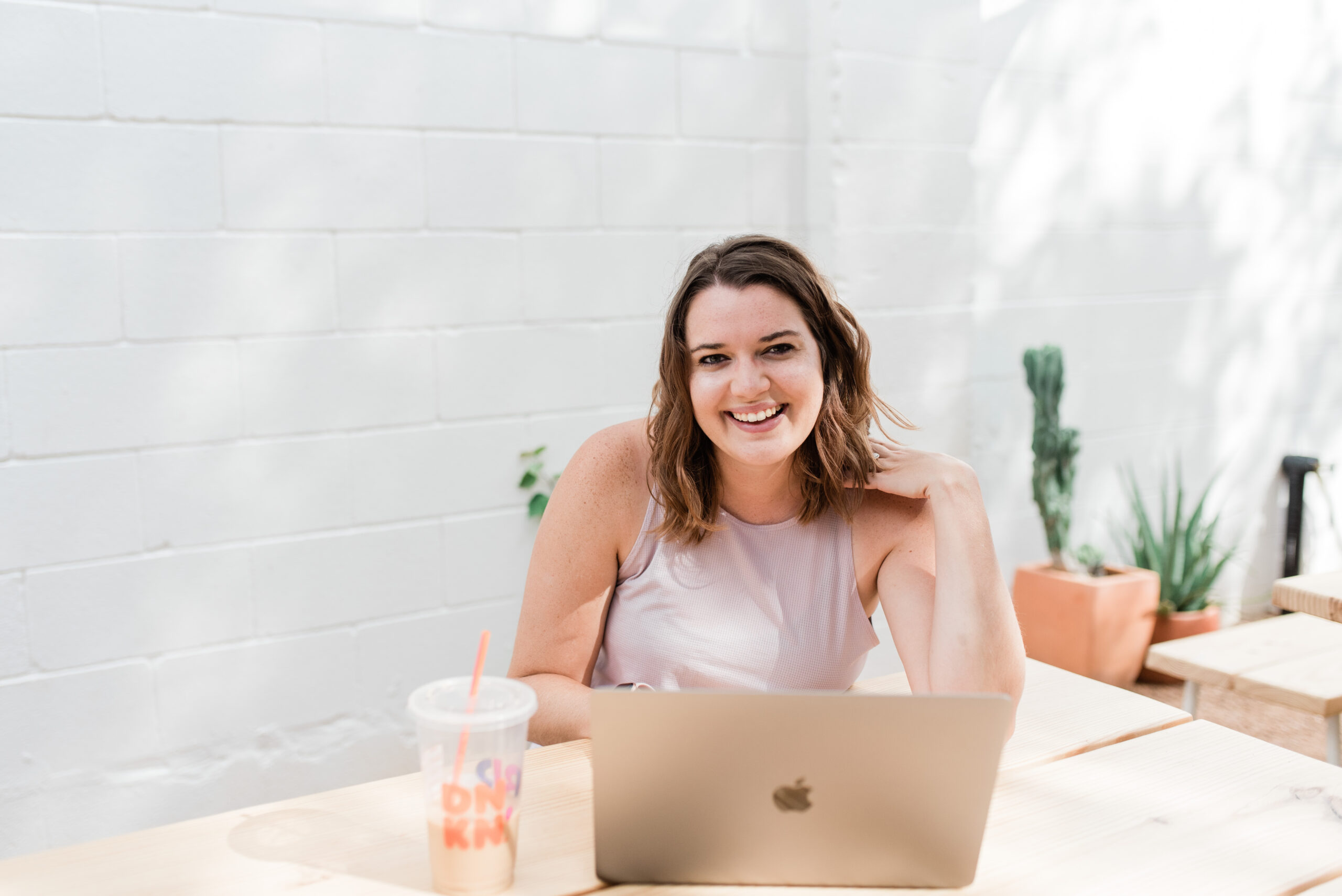 A person smiles while sitting at a table with a laptop and a drink in a bright, minimalist setting with potted plants, making notes on Amanda Shuman's marketing strategies.