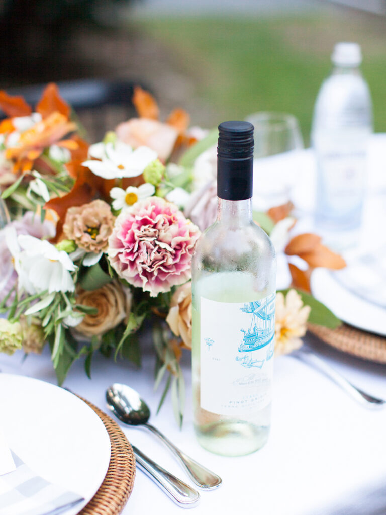 A bottle of white wine sits on an outdoor table set with flowers, plates, cutlery, and a water bottle, all perfectly arranged by a talented wedding planner.