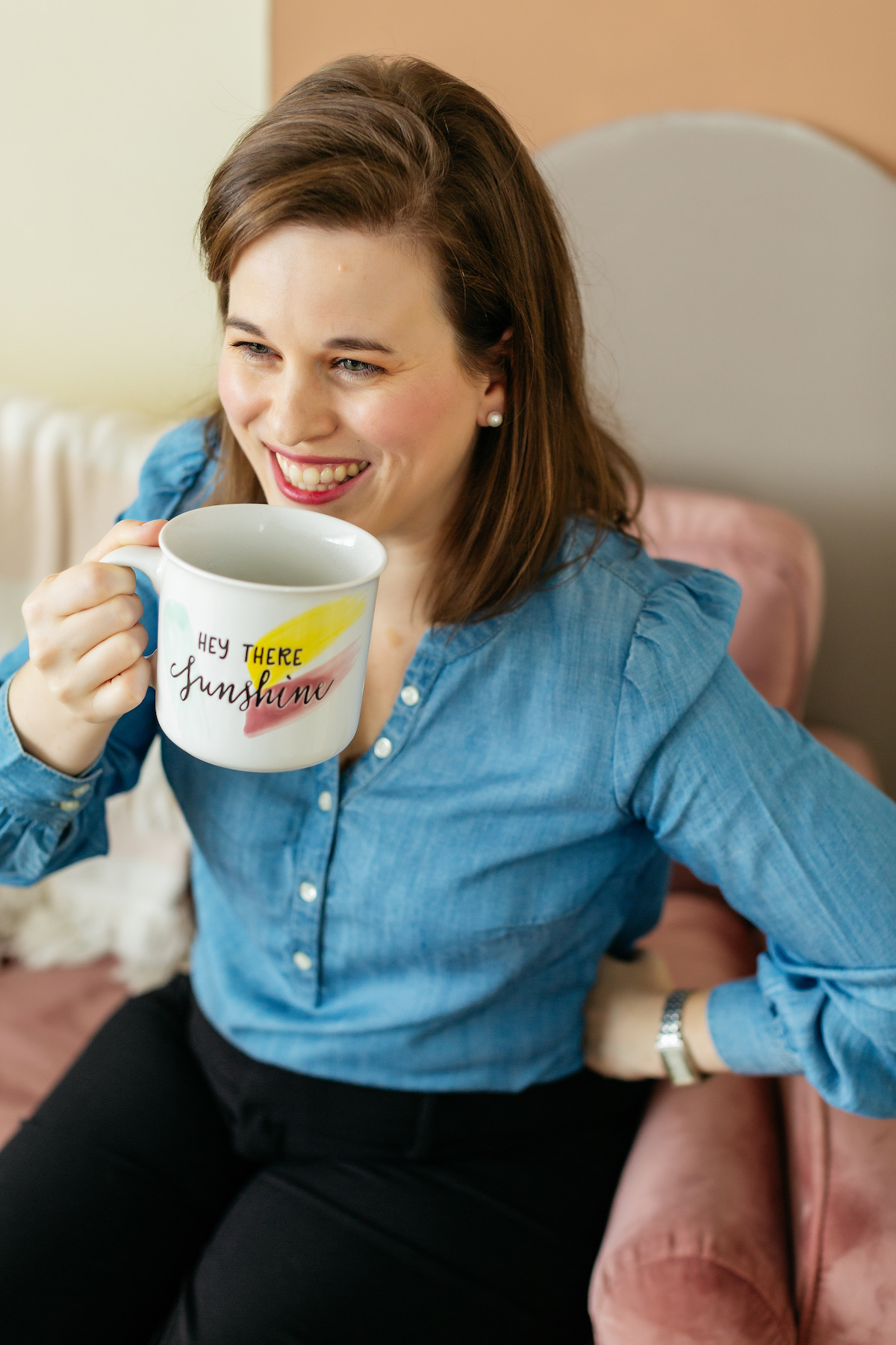 A woman in a denim shirt, sitting on a pink chair, enjoys her coffee in a mug that says "Hey There Sunshine," smiling as she plans new ways to boost local SEO for wedding pros.