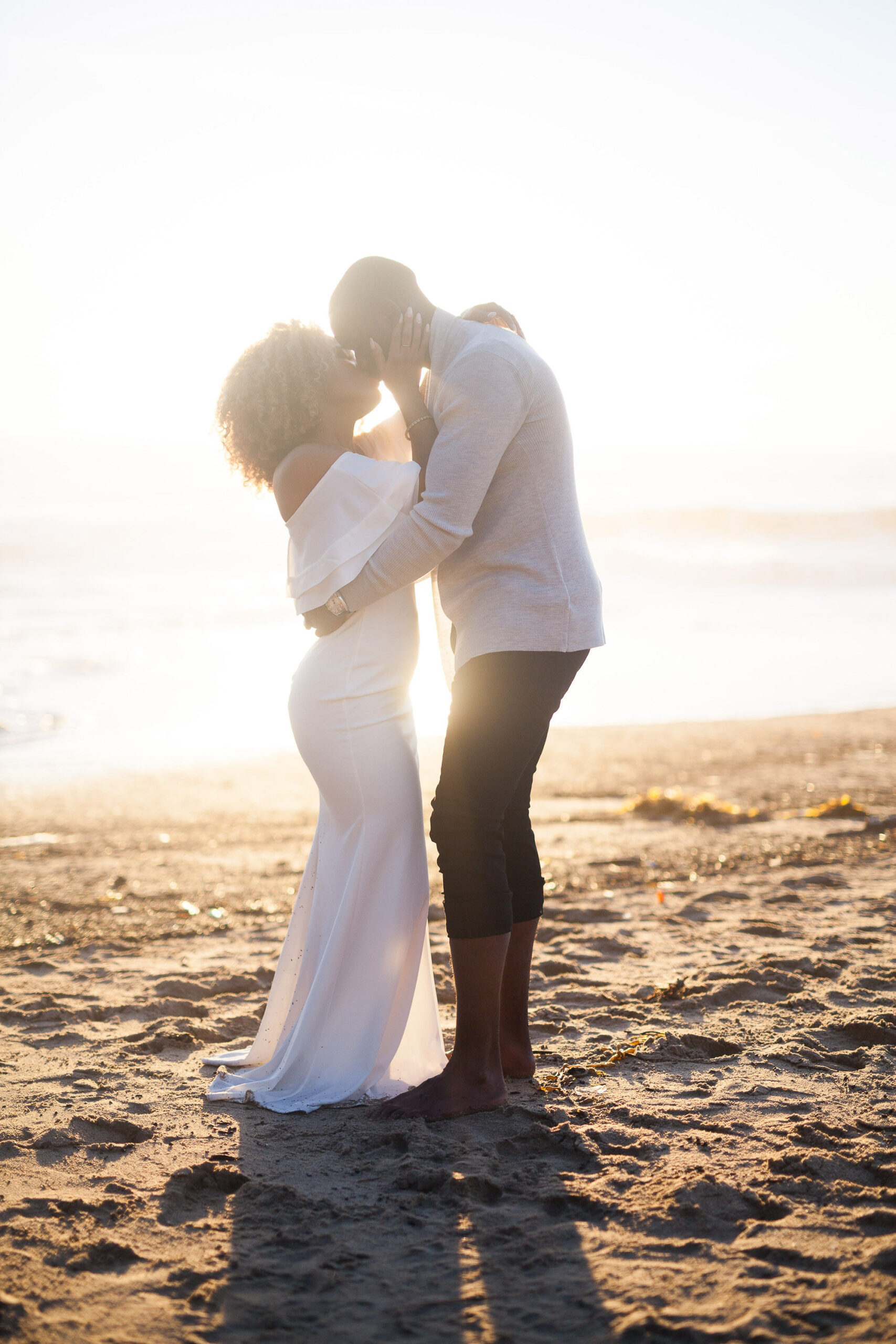 A couple embraces on a sandy beach at sunset, with soft light illuminating the scene. One in a white dress, the other in a light sweater and dark pants, captures the romance akin to a wedding sales process—elegantly choreographed and perfectly timed.