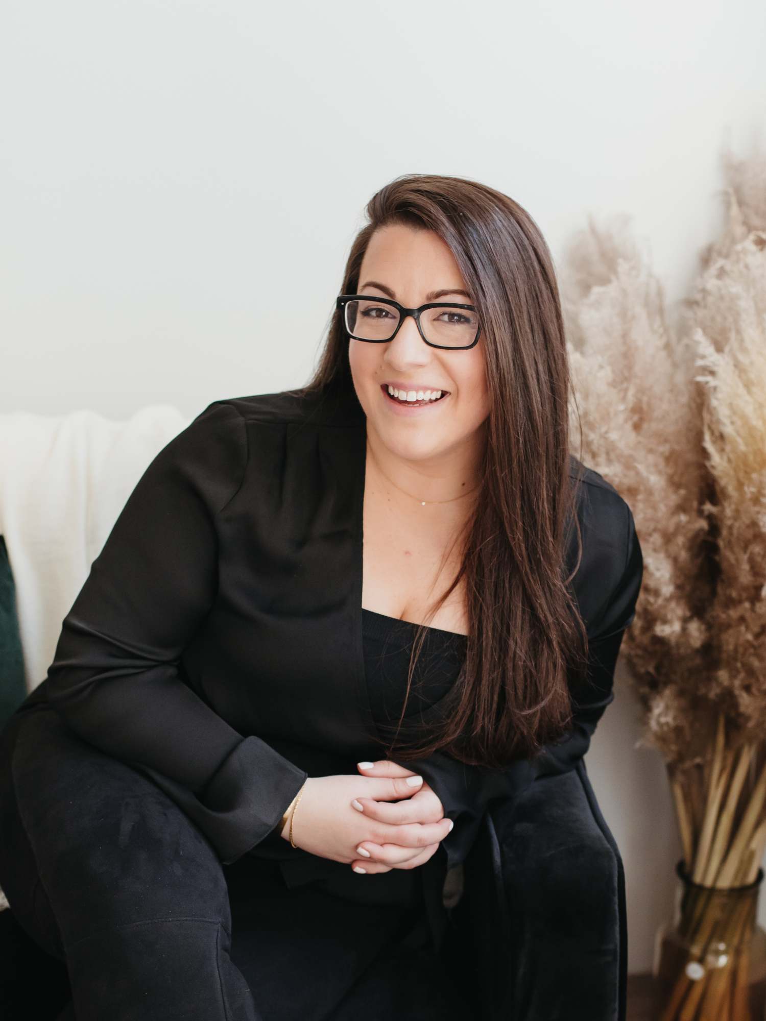 A person with long brown hair and glasses smiles while seated next to a bundle of dried pampas grass, contemplating ideas for client gifting for wedding pros.