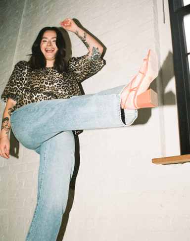 In a playful dance by the window, a person in leopard print top and light jeans joyfully lifts their leg, showcasing heels that are undeniably Madden Made.