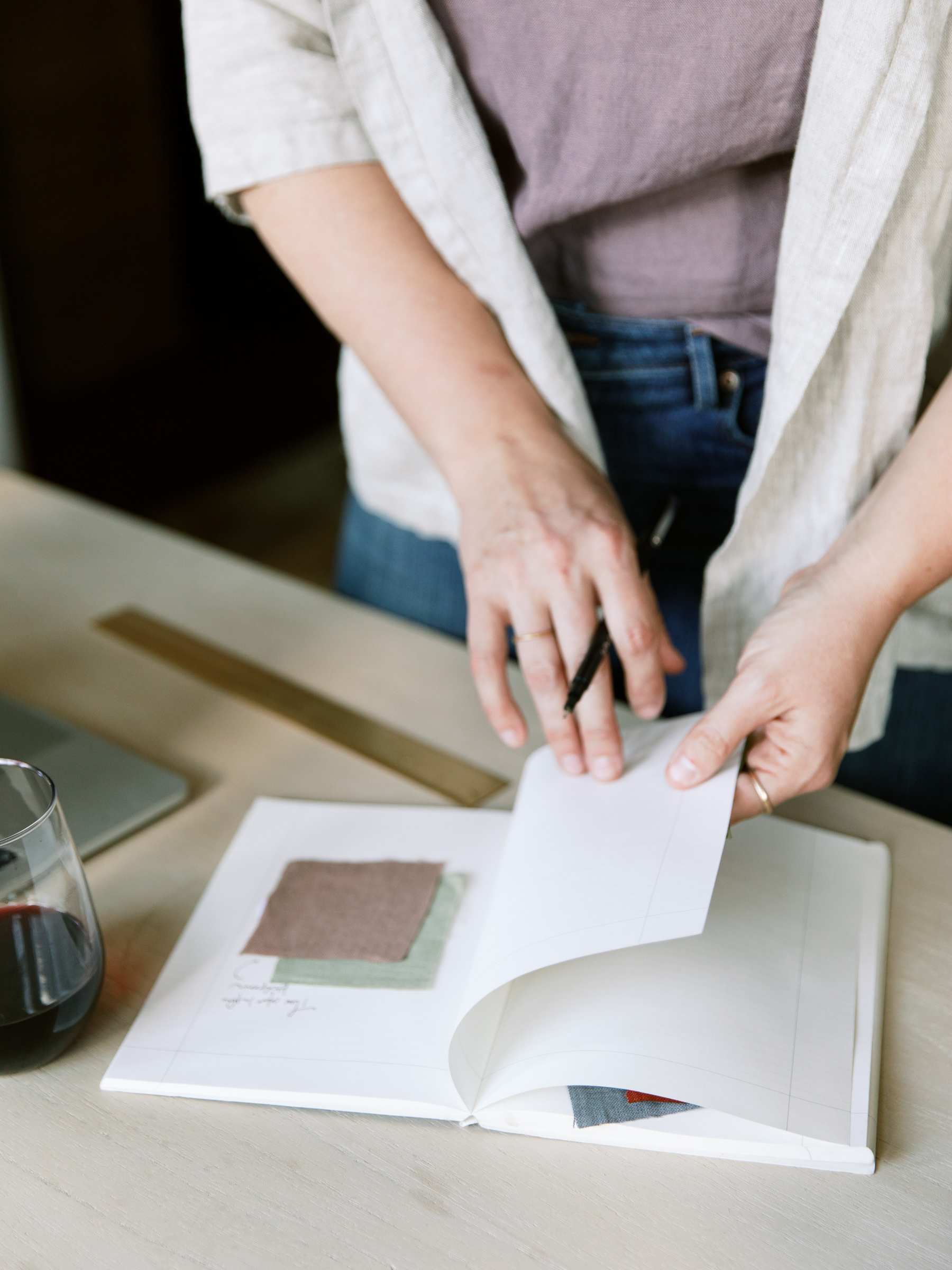 Person standing at a desk, expertly managing their time as they jot down notes in a notebook filled with fabric samples, alongside a laptop and a glass of red wine—an ideal setup for wedding pros balancing creativity and business.