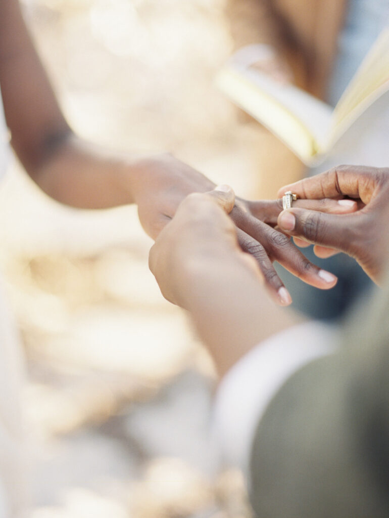 Close-up of a person placing a ring on another's finger in a natural outdoor setting, beautifully orchestrated by a full-service wedding planner.