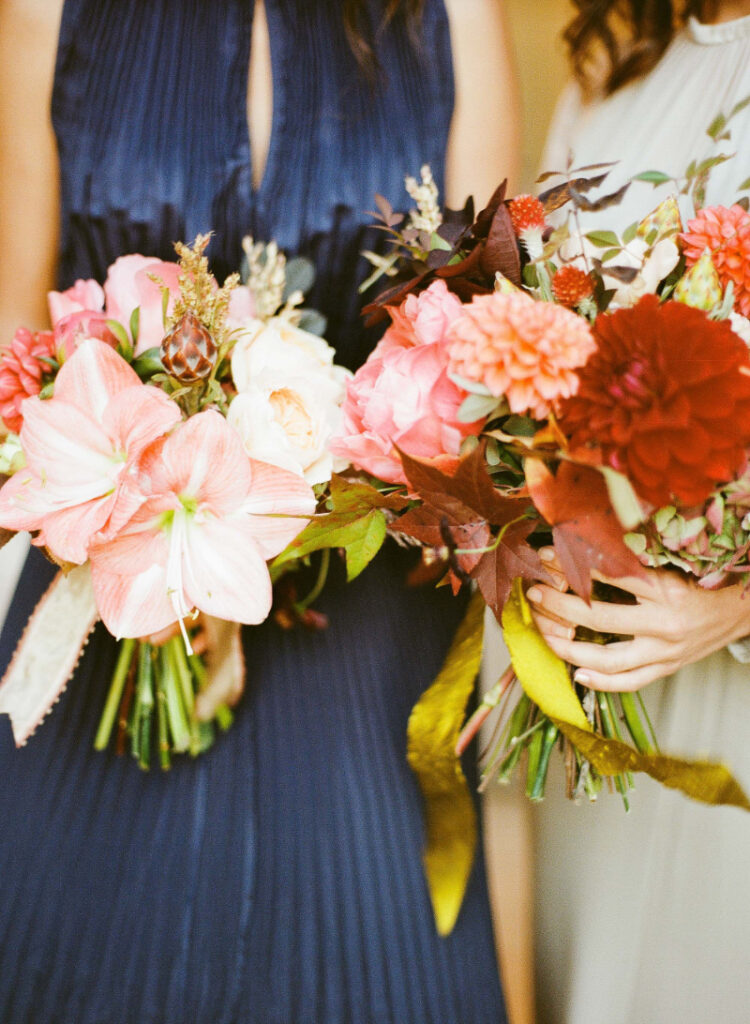 Two women holding colorful flower bouquets, perfect for a full-service wedding. The bouquets feature a vibrant mix of pink, red, and orange blooms with lush green and brown foliage. Both women are elegantly dressed in blue and beige dresses, embodying the art of wedding planning.