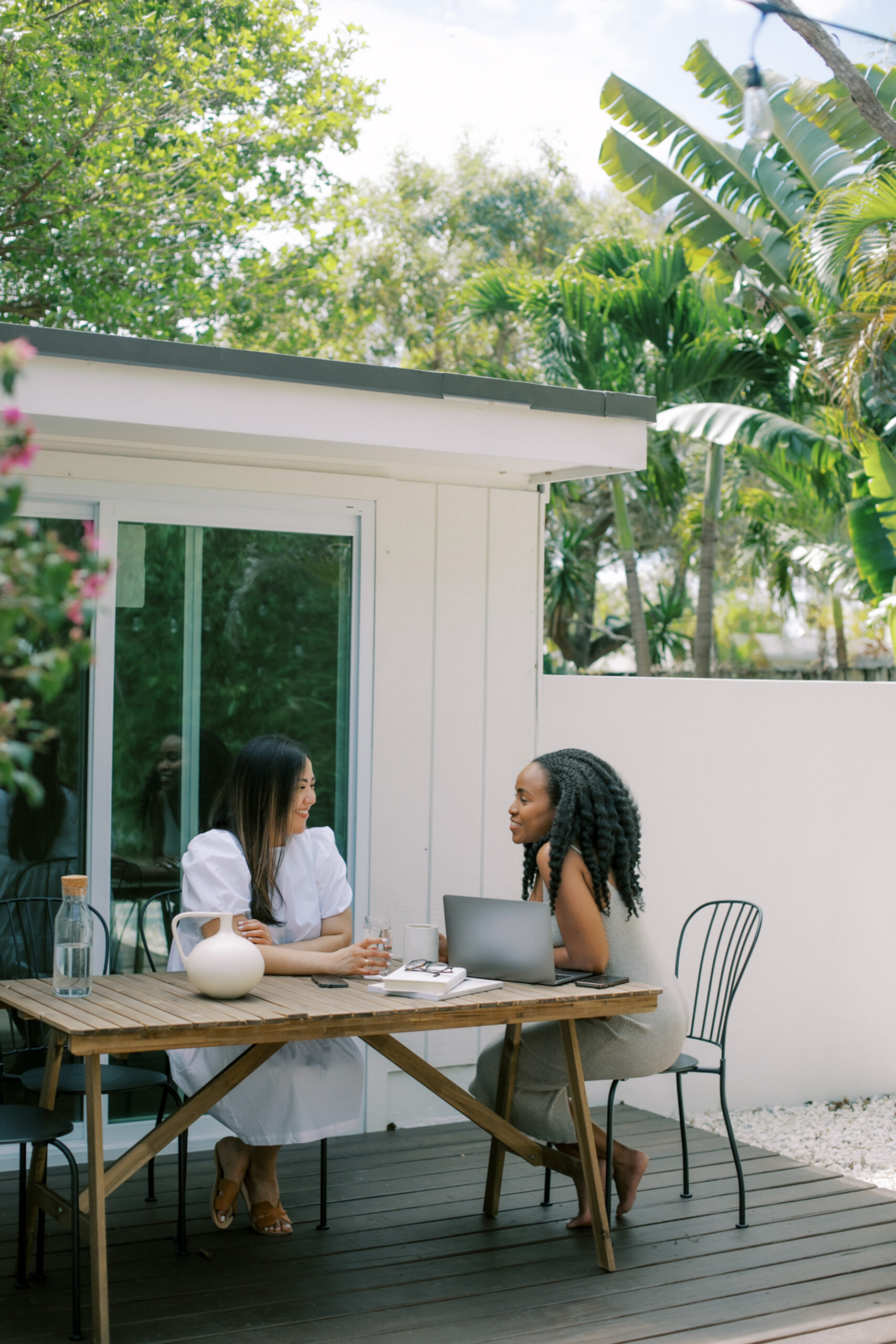 Two people sit at a wooden table on a patio, engrossed in conversation. A laptop and a vase serve as silent witnesses to their growing connection. Lush greenery surrounds them, creating an inviting setting where business meets friendship.