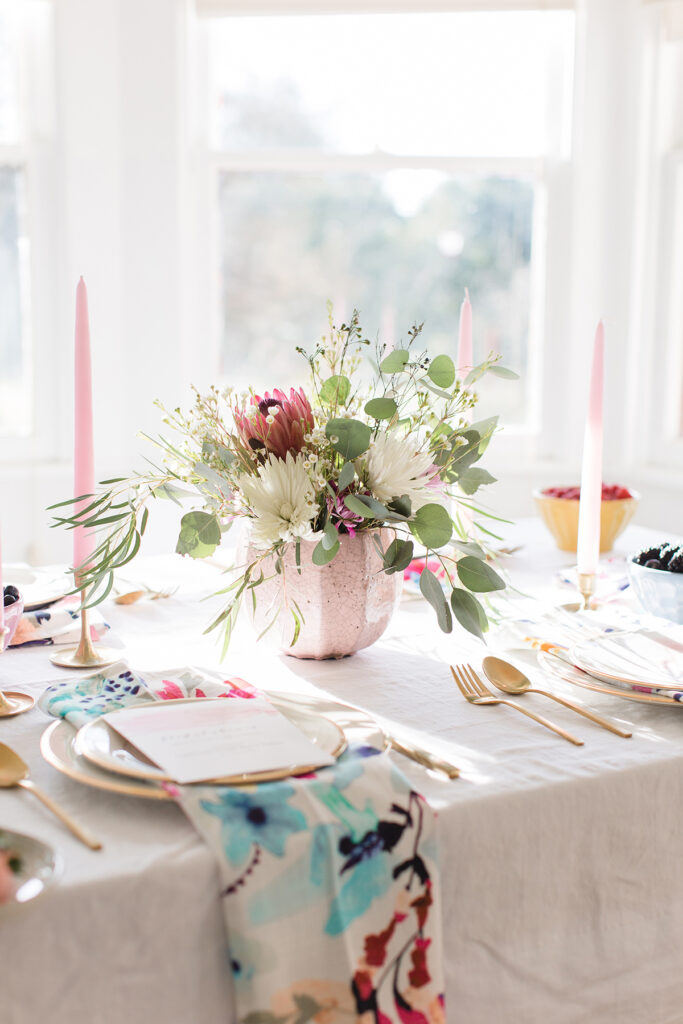 A bright dining table set with gold cutlery, pastel candles, floral centerpiece in a pink vase, and colorful napkins—perfect inspiration or blogging ideas for wedding planners—with natural light streaming through windows.