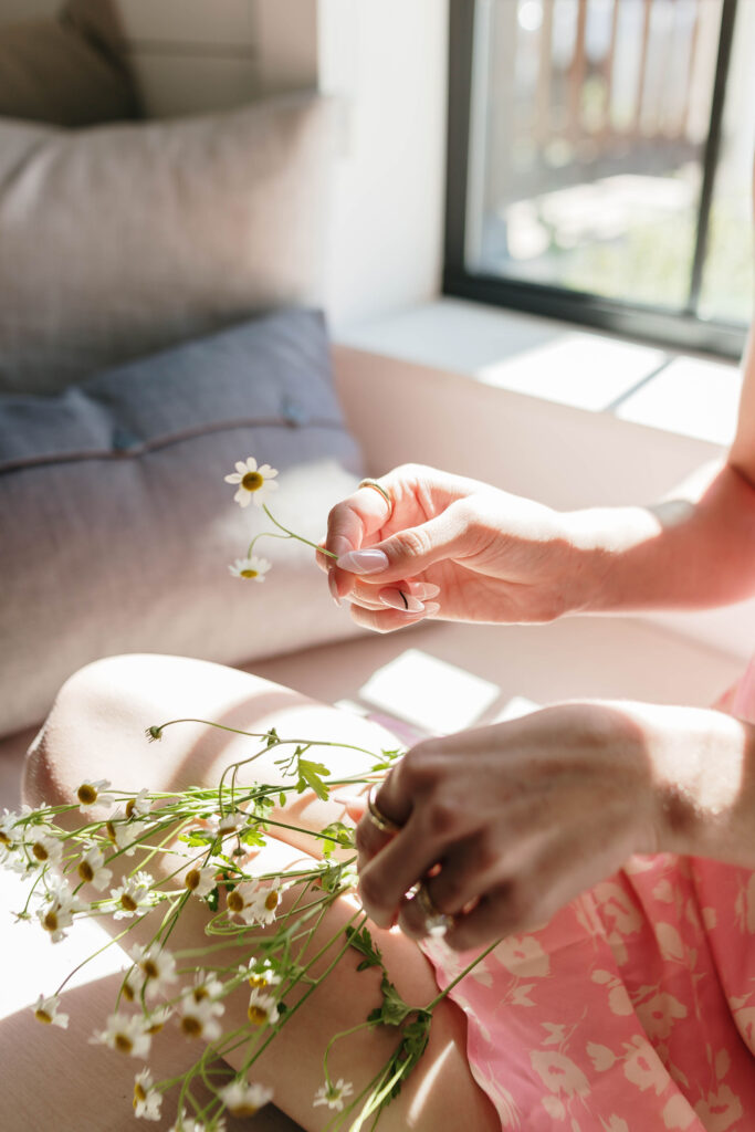 A person in a pink floral dress sits by a window, holding and picking petals off small daisies, perhaps pondering blogging ideas for wedding planners on a quiet afternoon.