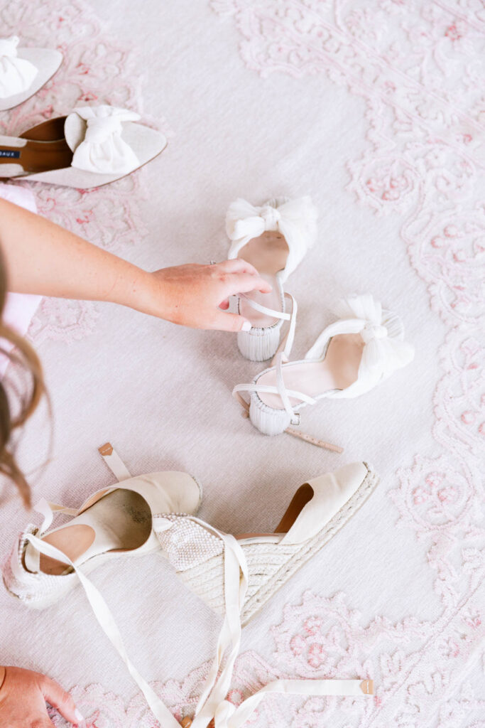 A person reaches for a pair of white heeled shoes with bows, surrounded by other white footwear on a light pink patterned rug—perfect for anyone looking to coordinate a wedding or seeking inspiration for wedding planning.