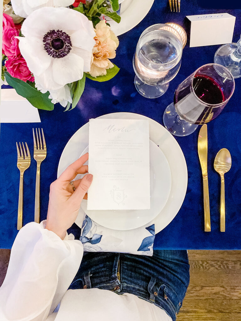 A person holds a menu over a white plate on a blue tablecloth, with gold cutlery, wine and water glasses, and a floral centerpiece—perfect inspiration for anyone looking to coordinate a wedding or elevate their wedding planning details.