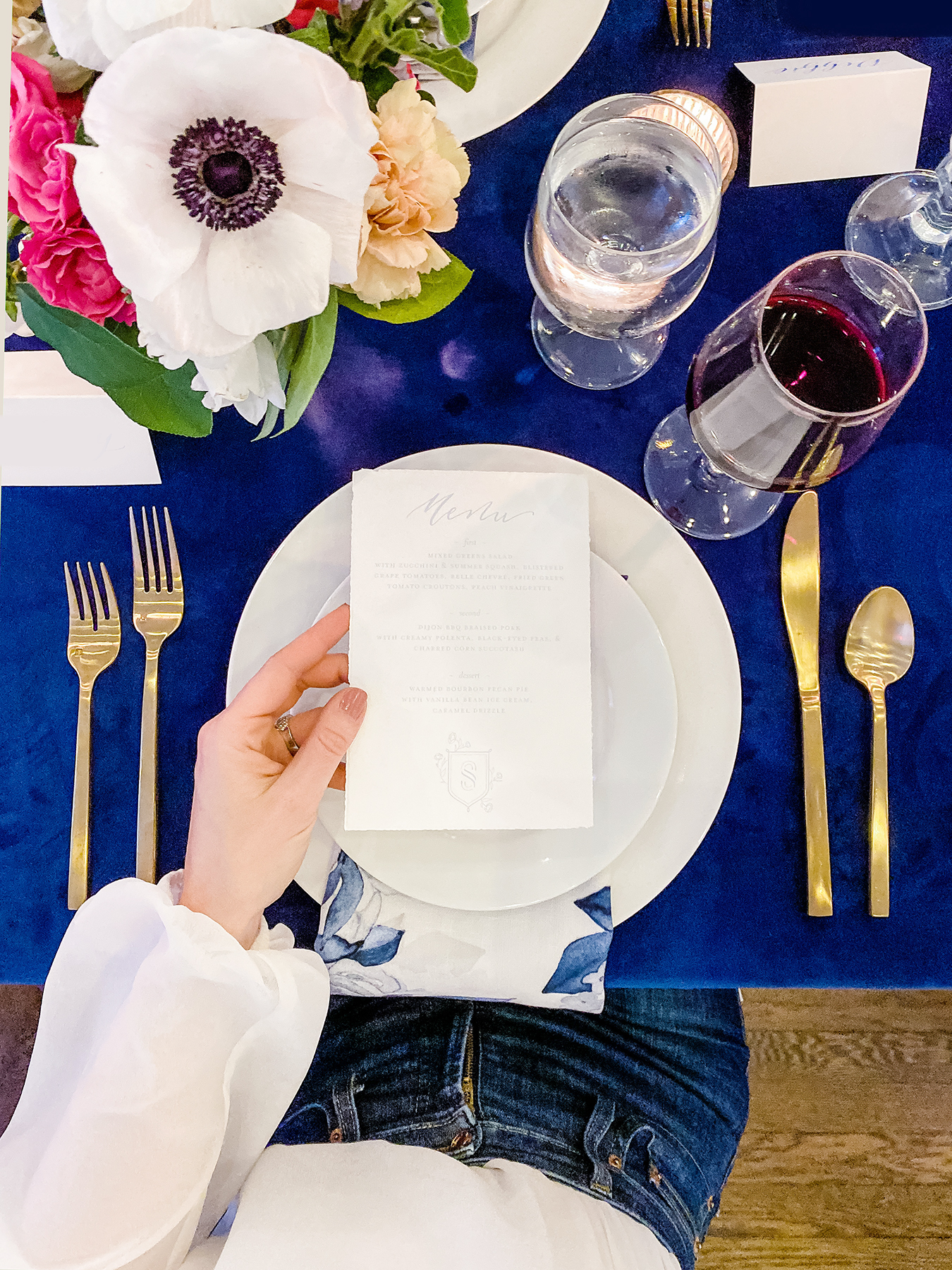 A person holds a menu over a white plate on a blue tablecloth, with gold cutlery, wine and water glasses, and a floral centerpiece—perfect inspiration for anyone looking to coordinate a wedding or elevate their wedding planning details.