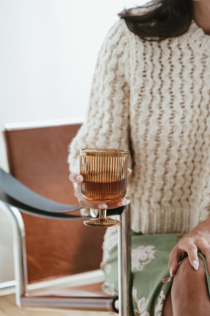 A person in a cream cable-knit sweater and floral skirt sits on a chair, holding an amber-colored ribbed glass&mdash;perfect for capturing candid moments or sharing wedding reviews after the celebration.