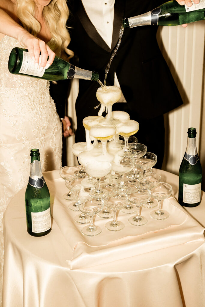 A couple in formal attire pours champagne into a tower of glasses on a satin-covered table, celebrating as they ask for wedding reviews to capture special memories from guests.