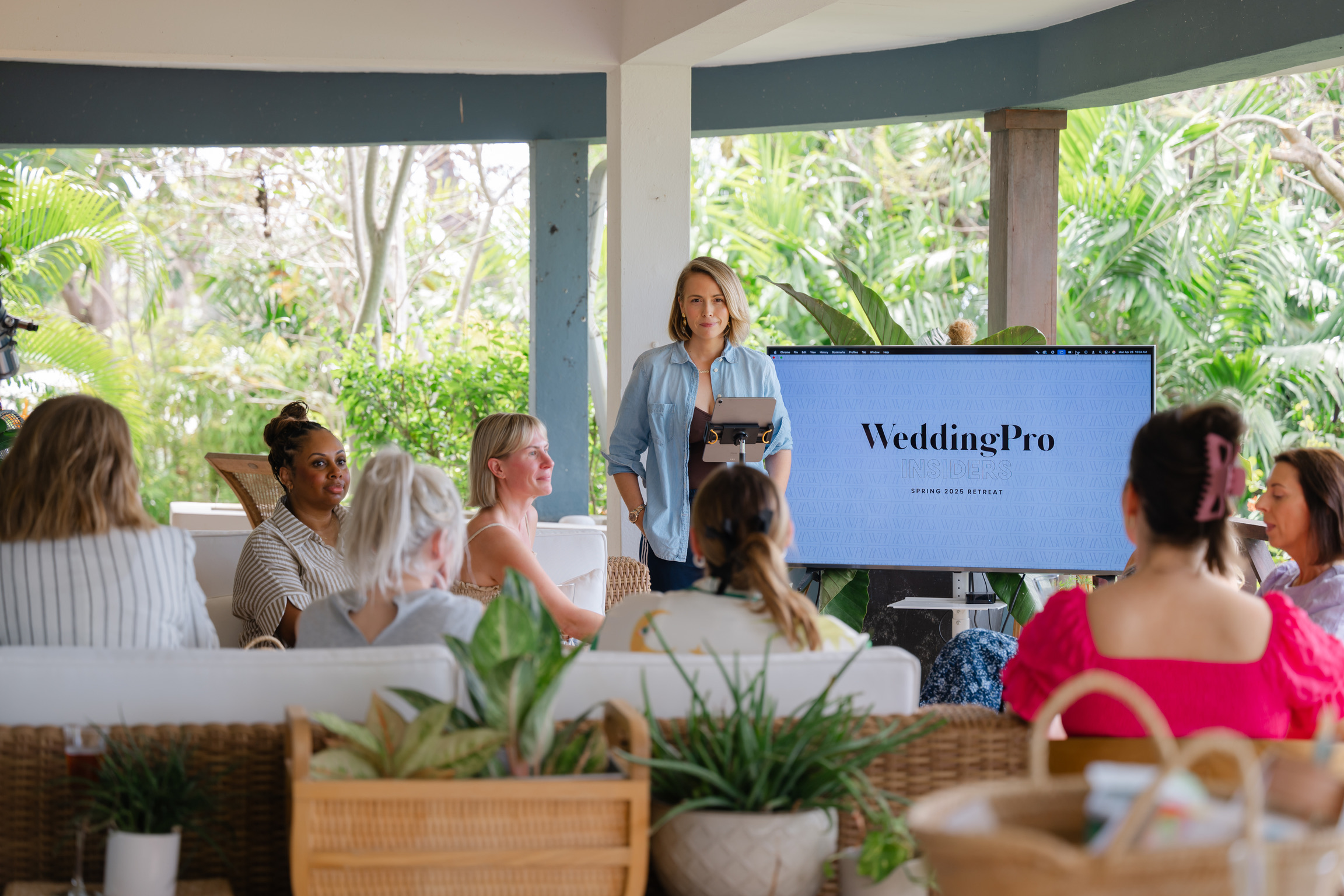 A woman stands at the front of a group, presenting a "WeddingPro" slideshow on a large screen to an audience seated in a bright, plant-filled room during a Wedding Business Retreat.