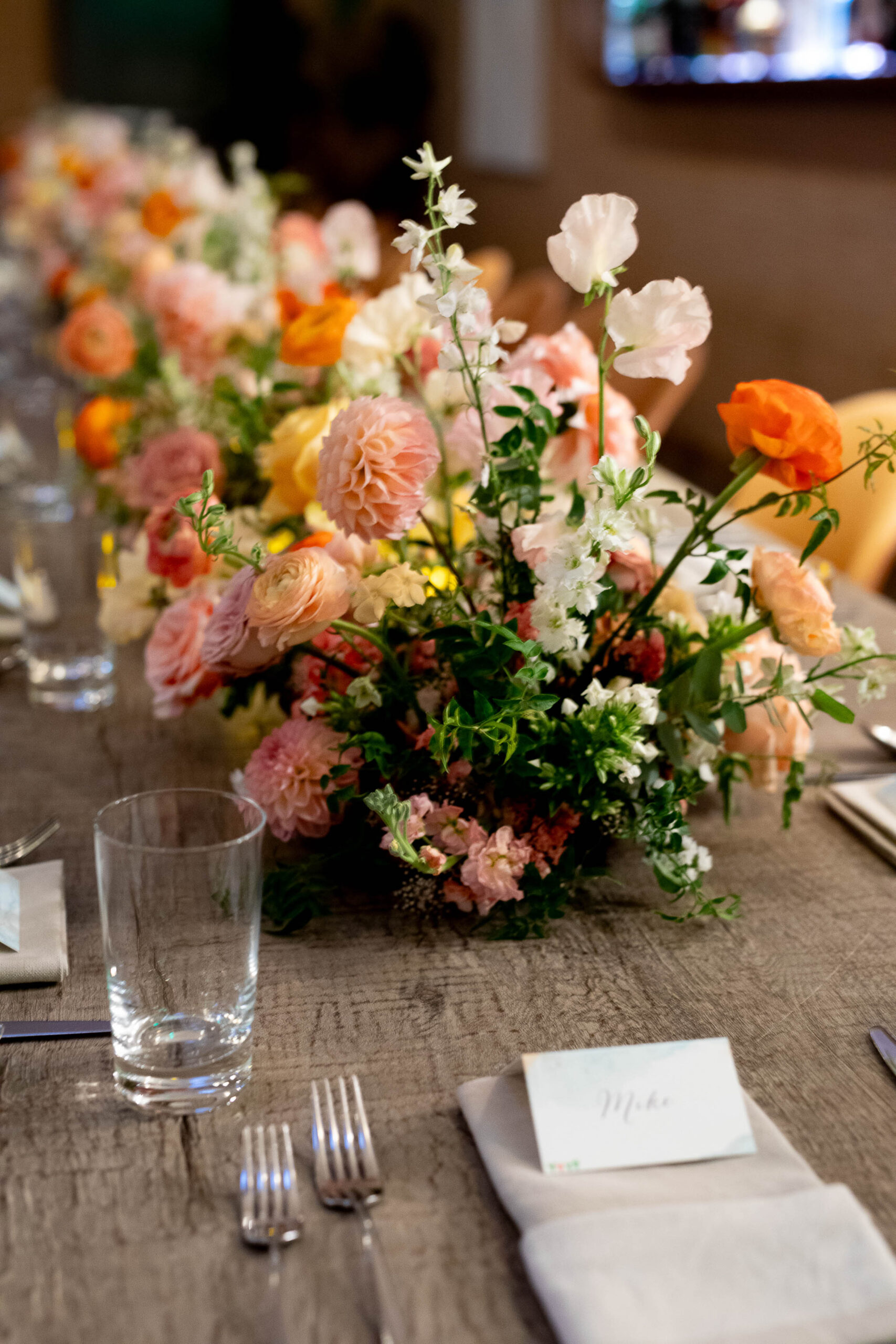 A long dining table set with clear glasses, cutlery, and napkins, featuring a vibrant centerpiece of assorted pink, yellow, and orange flowers—perfect inspiration for growing your wedding business.