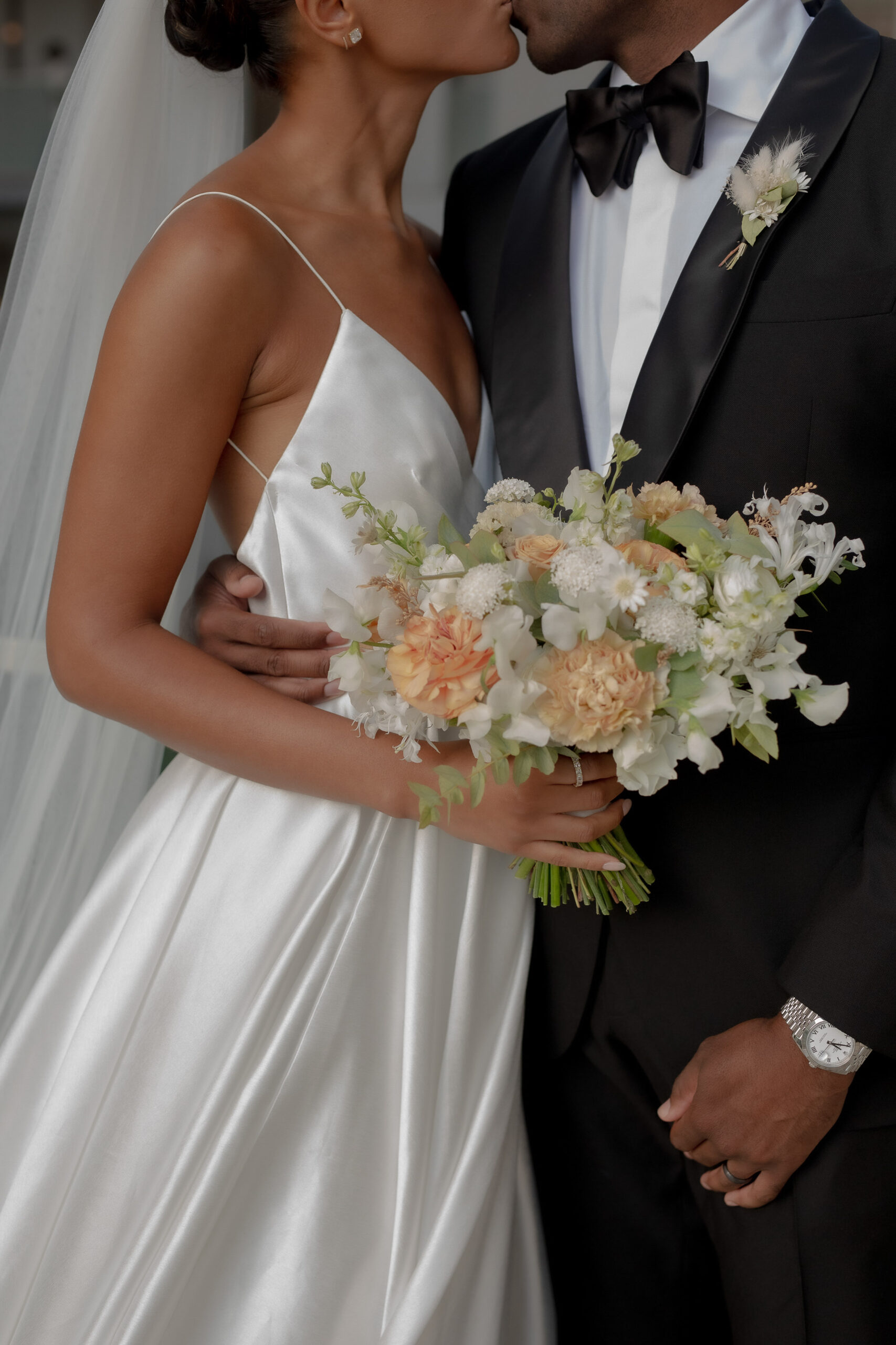 A bride in a white satin gown and a groom in a black tuxedo share a kiss; the bride holds a bouquet of peach and white flowers—a perfect moment for those growing your wedding business.