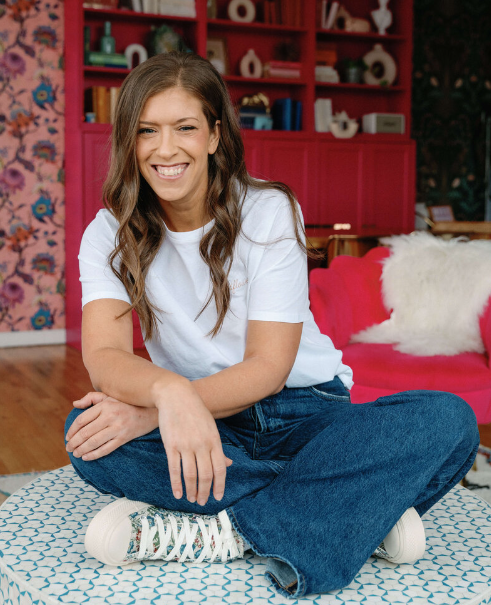 A woman with long brown hair sits cross-legged on a patterned ottoman, smiling in a colorful living room with red shelves and floral wallpaper—a behind the brand glimpse from Wildflower Events' creative event design space.