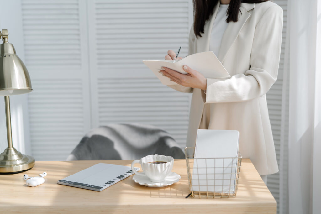 A woman in a white blazer stands at a desk, writing personal development goals on a notepad. The desk features a lamp, documents, cup and saucer, wire basket, and a pair of wireless earbuds.