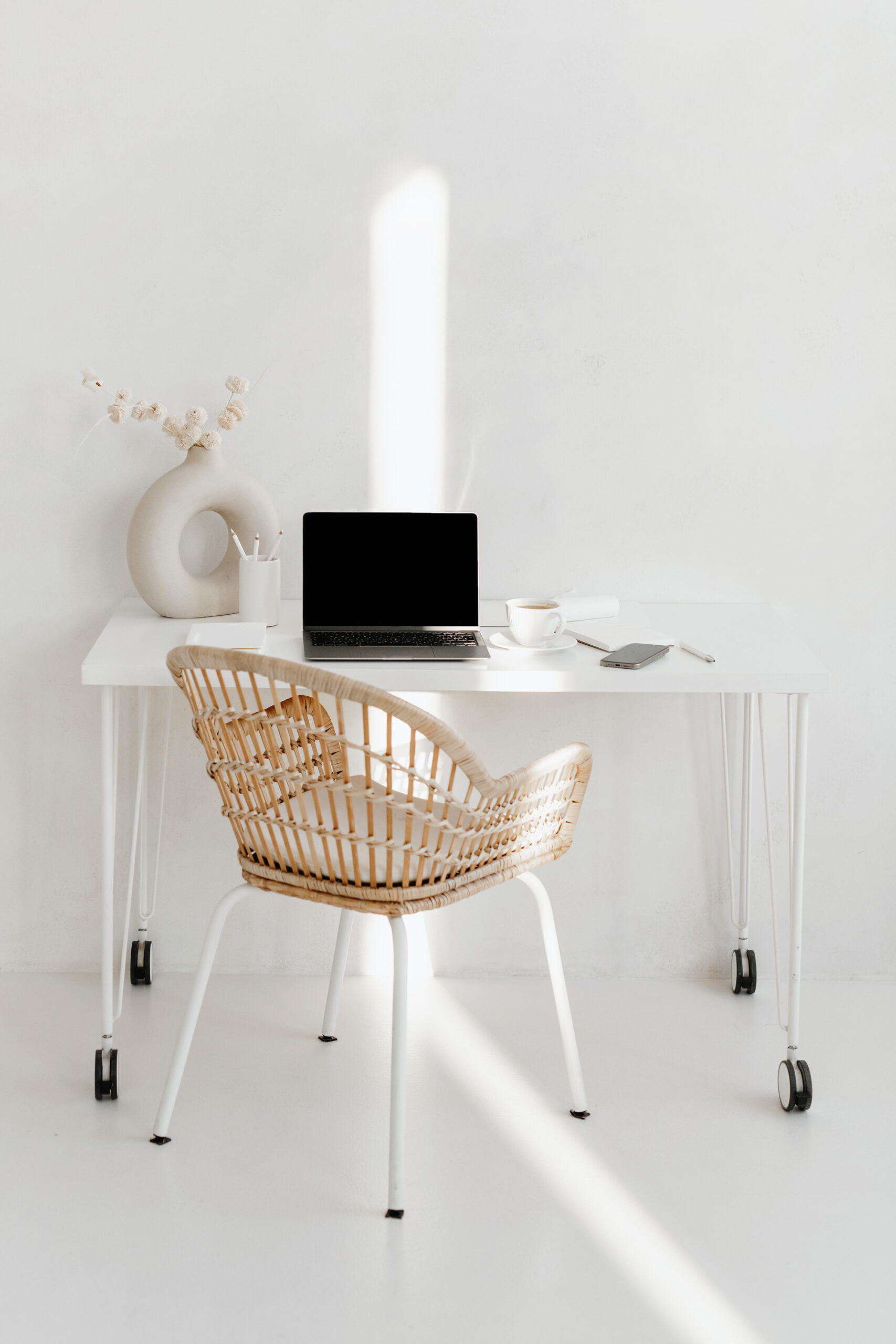 A minimalist workspace with a white desk, laptop, cup, notebook for personal development goals, pen holder, vase with dried flowers, and a wicker chair in a bright room.