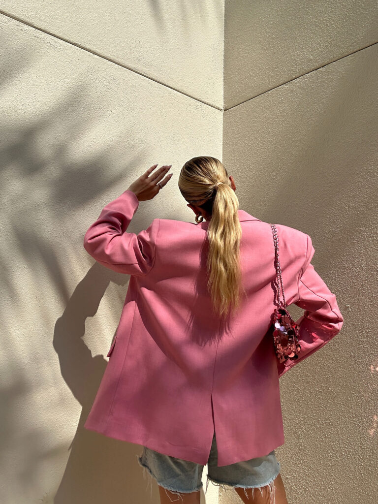A person with long blond hair in a ponytail, wearing a pink blazer and denim shorts, stands facing a beige wall with one hand raised and a pink purse over their shoulder—capturing the essence of building a personalized brand.