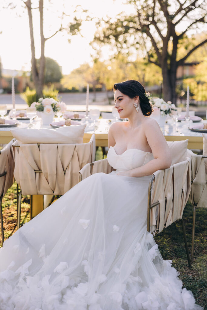 A woman in a strapless white wedding dress sits at an outdoor table decorated with flowers and candles, sunlight filtering through the trees—the perfect setting for celebrating with wedding weekend planning packages.