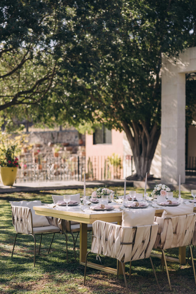 A decorated outdoor dining table with six cushioned chairs, floral centerpieces, and candles is set under a large tree in a garden, perfect for gatherings or elegant affairs like wedding weekend planning packages.