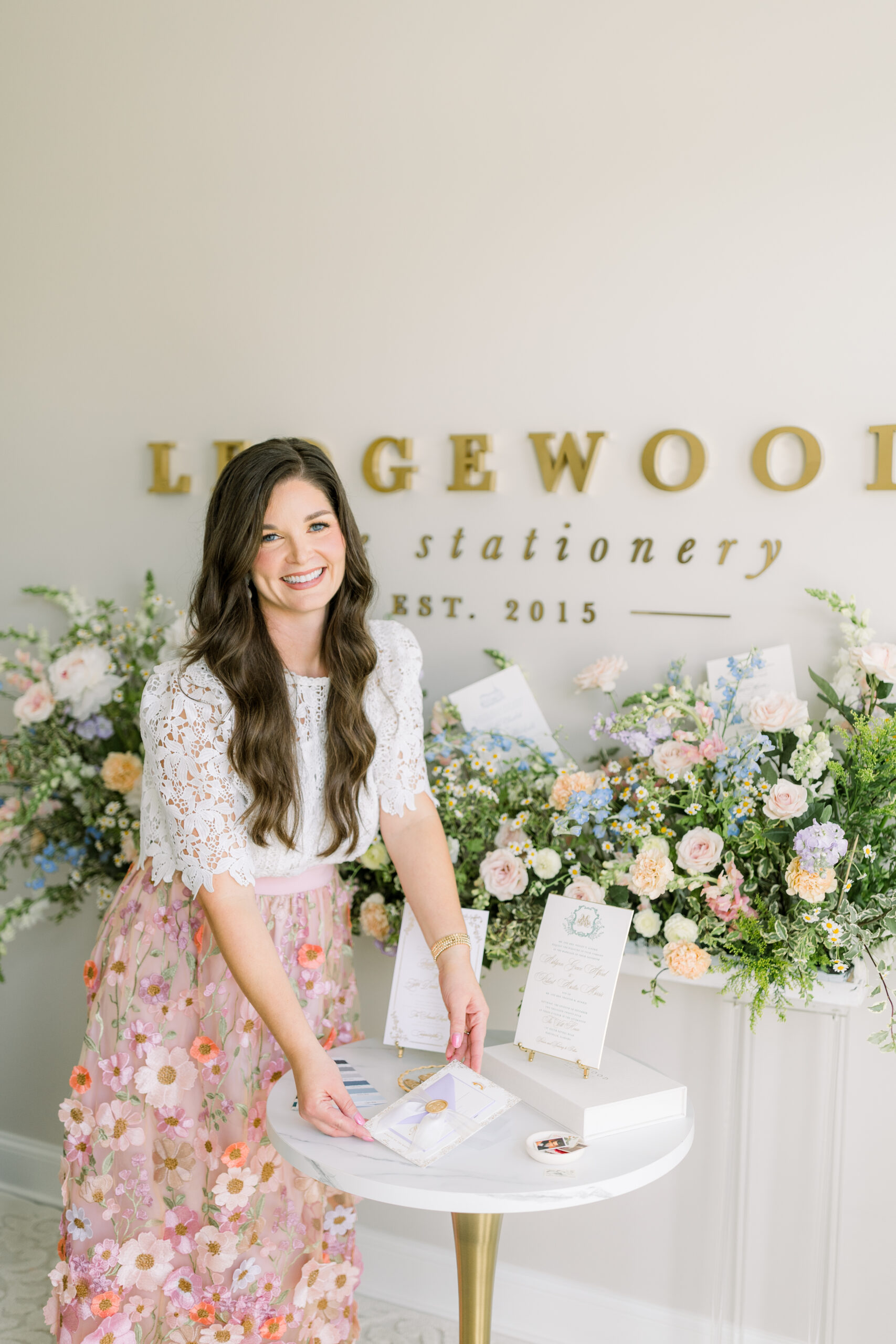 A woman stands at a small round table arranging stationery items, with floral arrangements and a "Ledgewood Fine Stationery Est. 2015" sign in the background.