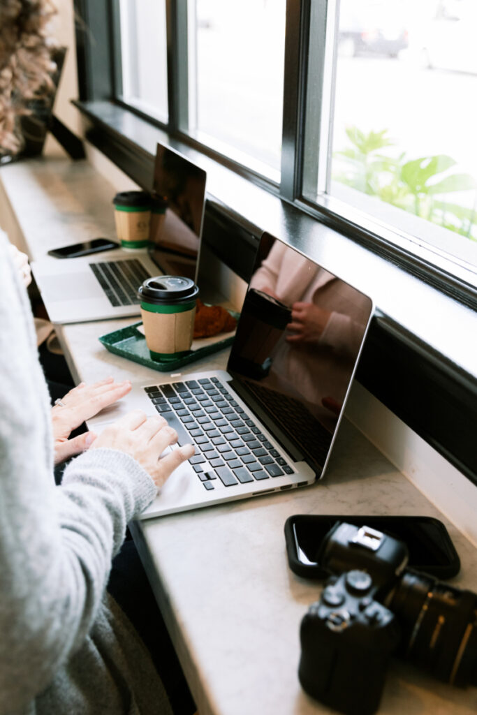 Person typing on a laptop at a cafe counter with coffee, croissant, phone, and camera nearby, seated by a large window.