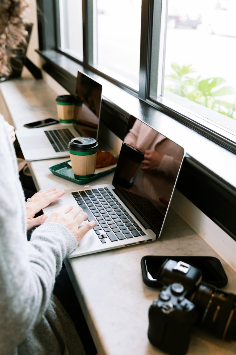Person typing on a laptop at a cafe counter with coffee, croissant, phone, and camera nearby, seated by a large window.