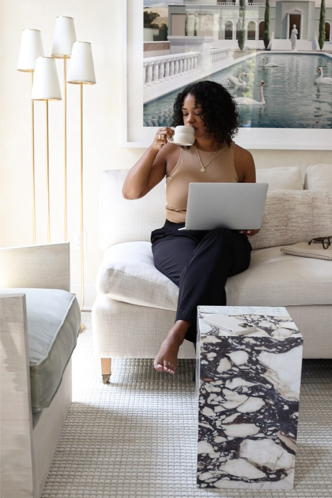 Woman sitting on a couch with a laptop, sipping coffee, modern lamp and framed artwork in the background.