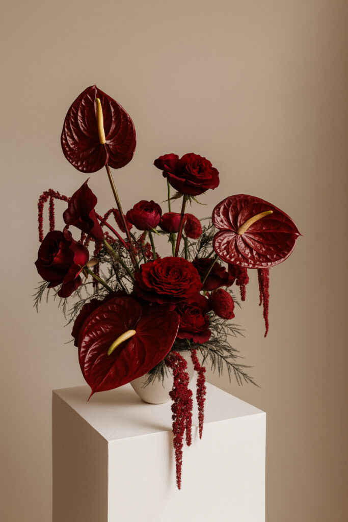 A sculptural floral arrangement on a white pedestal styled as dark romantic wedding decor, featuring deep burgundy anthuriums, velvety roses, cascading amaranthus, and textured greenery against a softly lit neutral background.