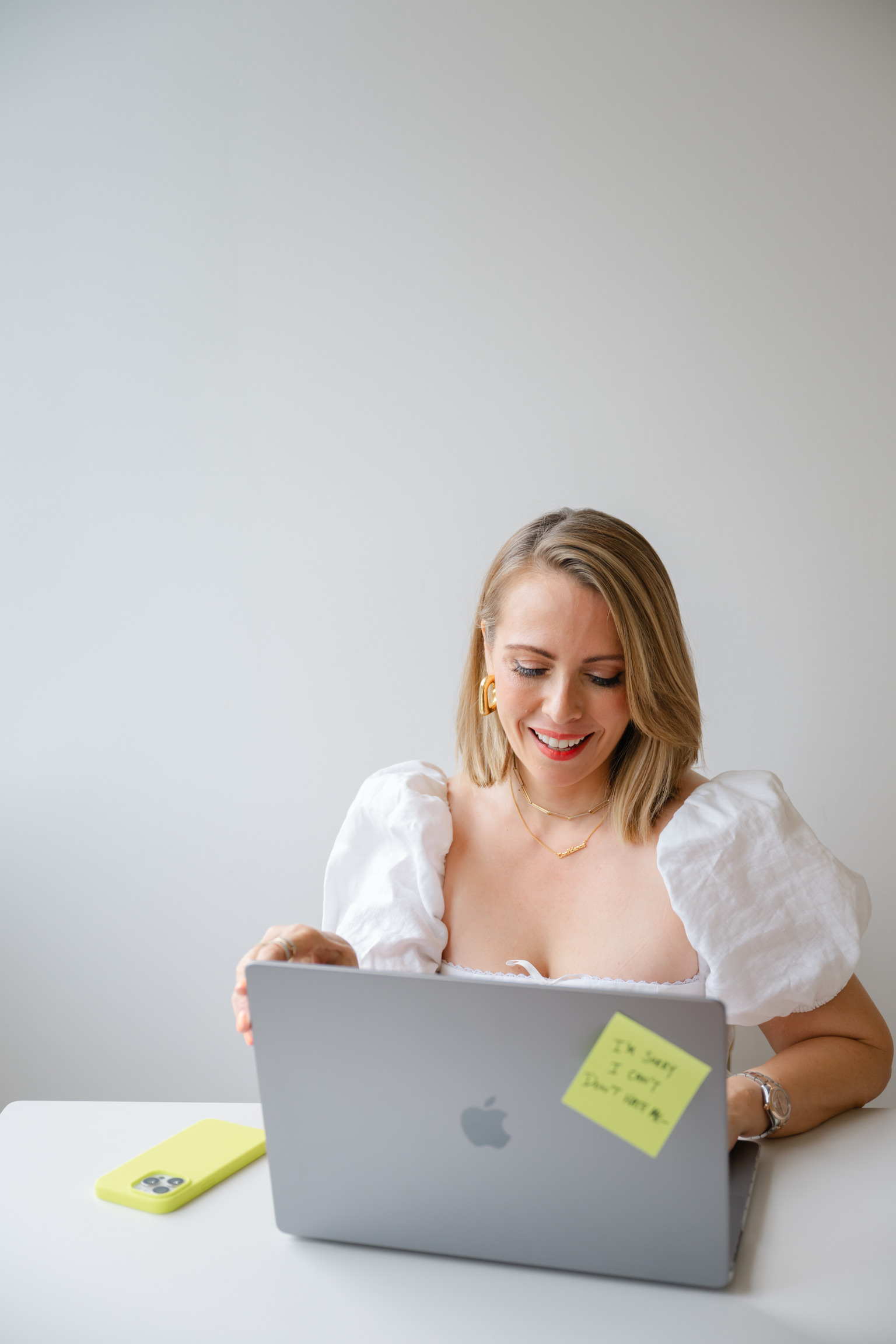 A woman in a white top sits at a desk, smiling at her laptop—perhaps managing her wedding business. A yellow sticky note is attached to the laptop, and a yellow phone case is on the desk.