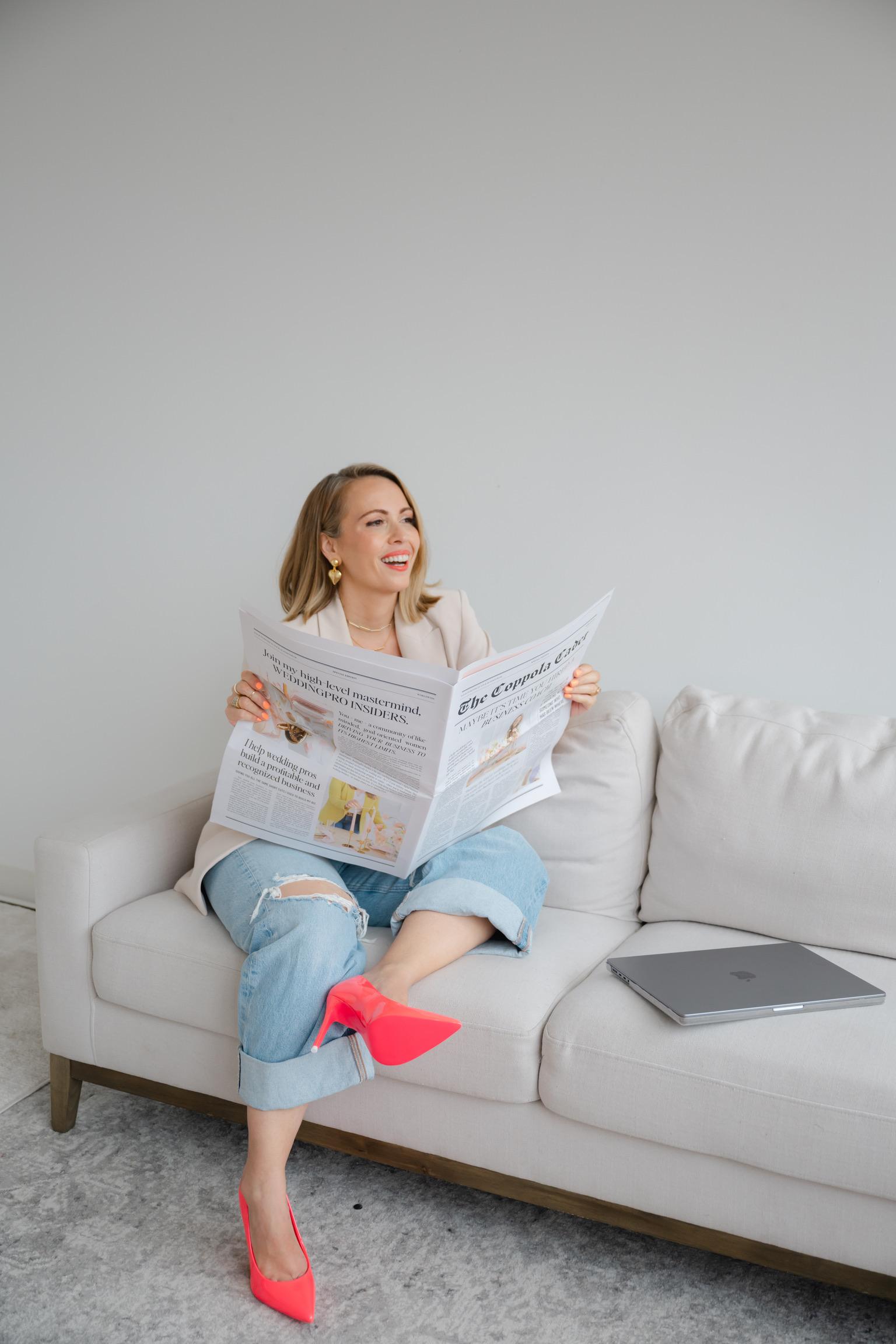 A woman sits on a white sofa, smiling while reading a newspaper article about wedding prices. She is wearing light blue jeans and bright pink shoes. A closed laptop rests on the sofa beside her.