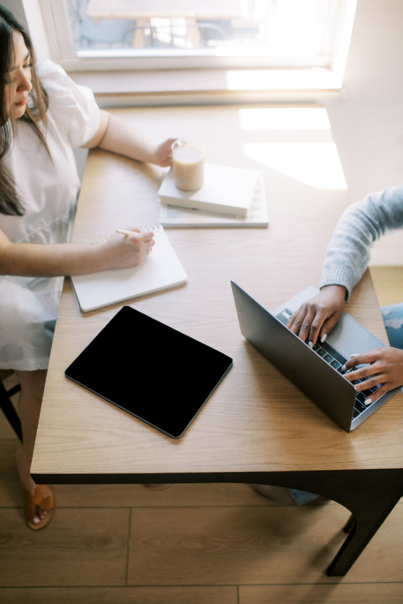 Two people work at a wooden table; one writes in a notebook with a coffee nearby, while the other types on a laptop, possibly searching how to train ChatGPT to write like you. A tablet rests on the table as daylight streams through a window.