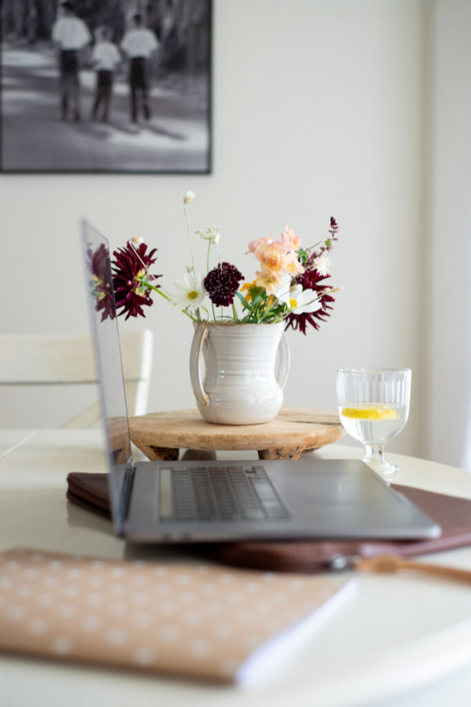 A laptop on a table next to a notebook, a glass of water with lemon, and a vase of assorted flowers—perfect for researching how to train ChatGPT to write like you; a black-and-white photo hangs on the wall in the background.
