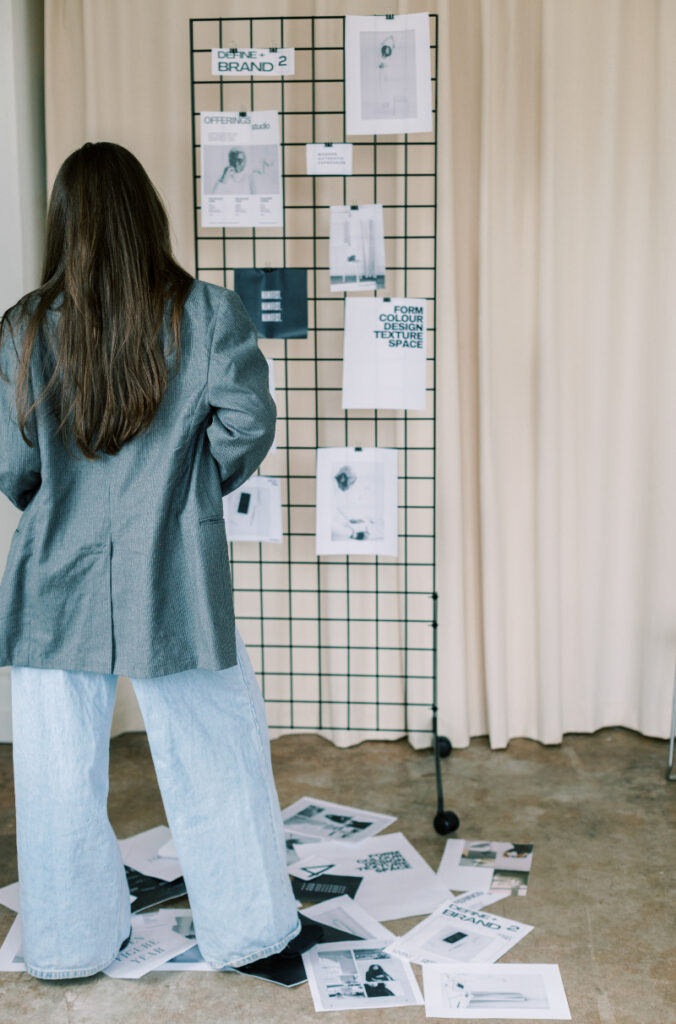 A person in a gray blazer and wide jeans stands before a wire board pinned with papers, as if brainstorming how to train ChatGPT to write like you—more notes are scattered across the floor.
