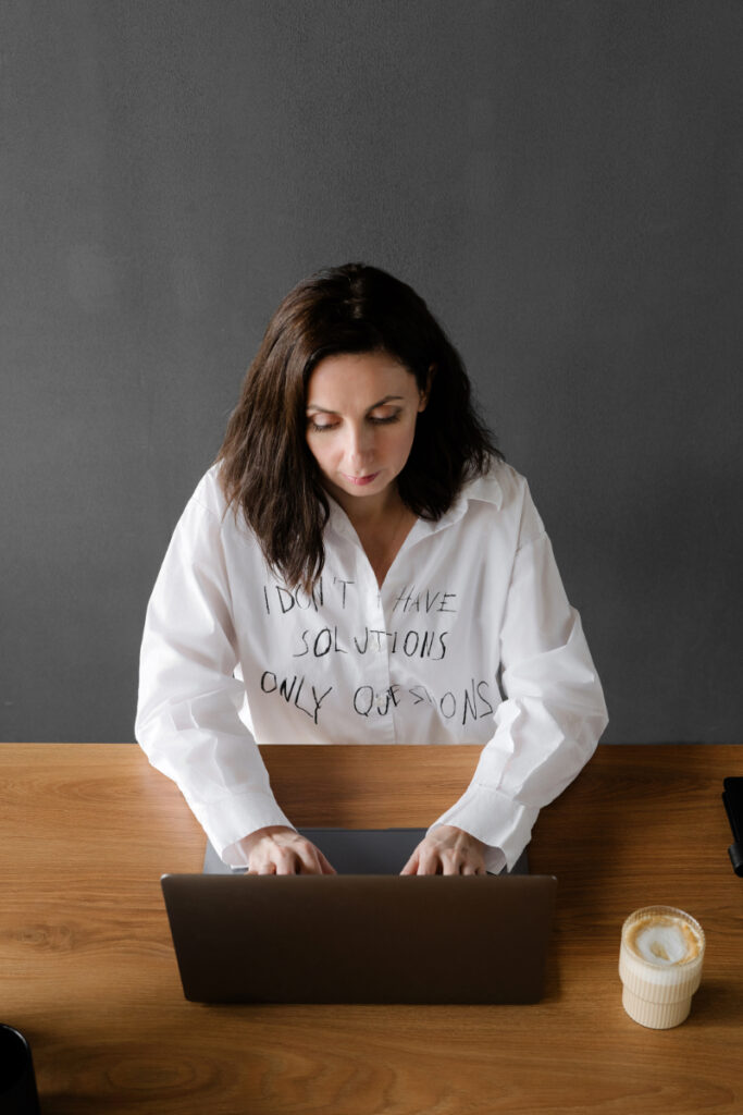A woman with dark hair types on a laptop at a wooden table, coffee nearby, wearing a white shirt that says "I don't have solutions, only questions," as she researches how to use ChatGPT for your wedding business.