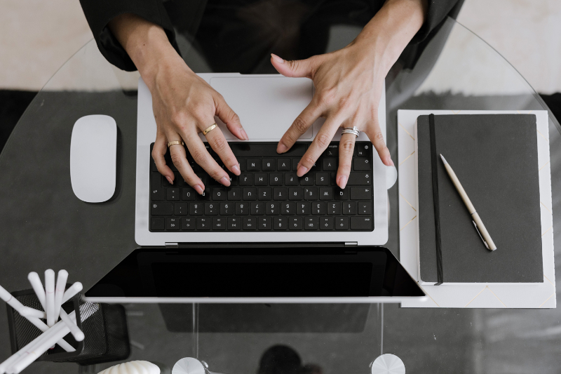 Person typing on a laptop at a glass desk with a notebook, pen, and computer mouse nearby—researching how to use ChatGPT for your wedding business.
