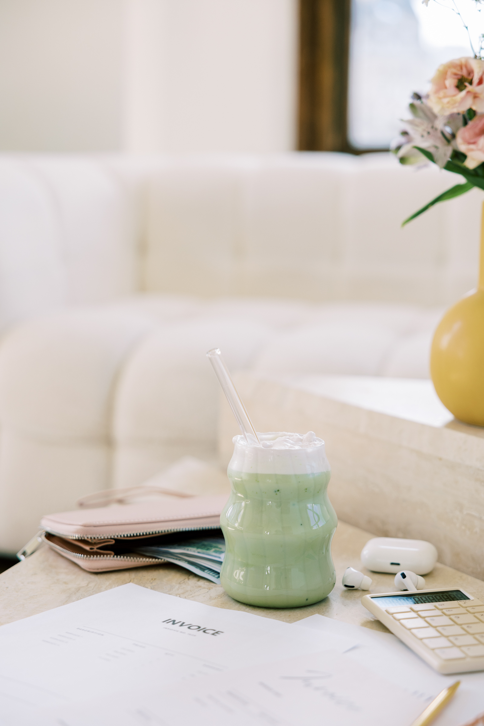 A green matcha latte with foam and a glass straw sits on a table next to paperwork, a calculator, wallet, AirPods case, and a yellow vase—ideal for wedding business planning or spotting red flags in contracts in a bright room.