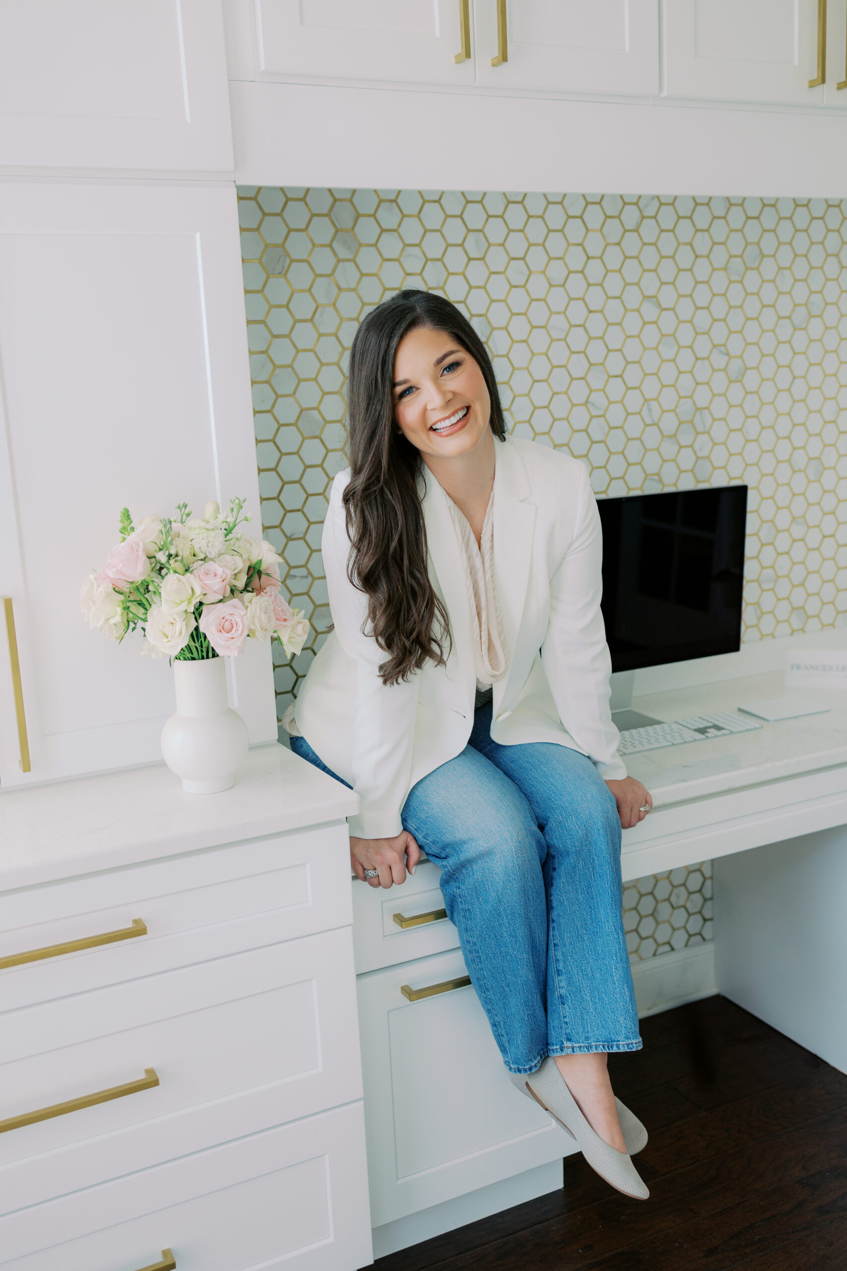 Woman in a white blazer and jeans sits on a white desk in a modern office with gold-accented cabinets, a vase of flowers, Ledgewood Fine Stationery, and a computer monitor.