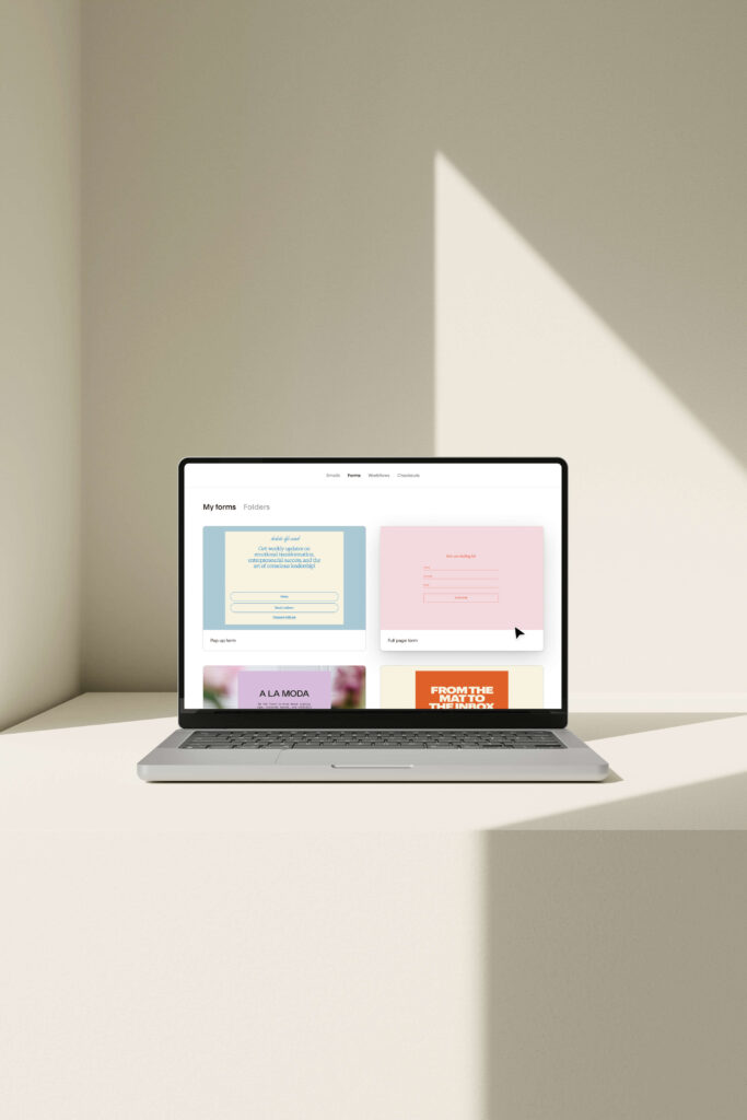 A laptop on a beige surface displays a Flodesk website with pastel-colored cards and navigation tabs, perfect for small businesses, under bright natural lighting.