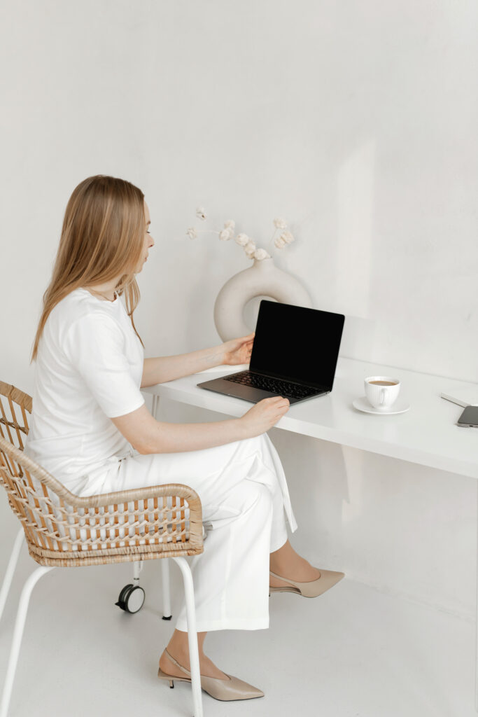 A woman in white clothing sits on a wicker chair at a white desk, using her laptop to browse a website platform for wedding planners. A cup on a saucer and a vase with dried flowers decorate the serene workspace.