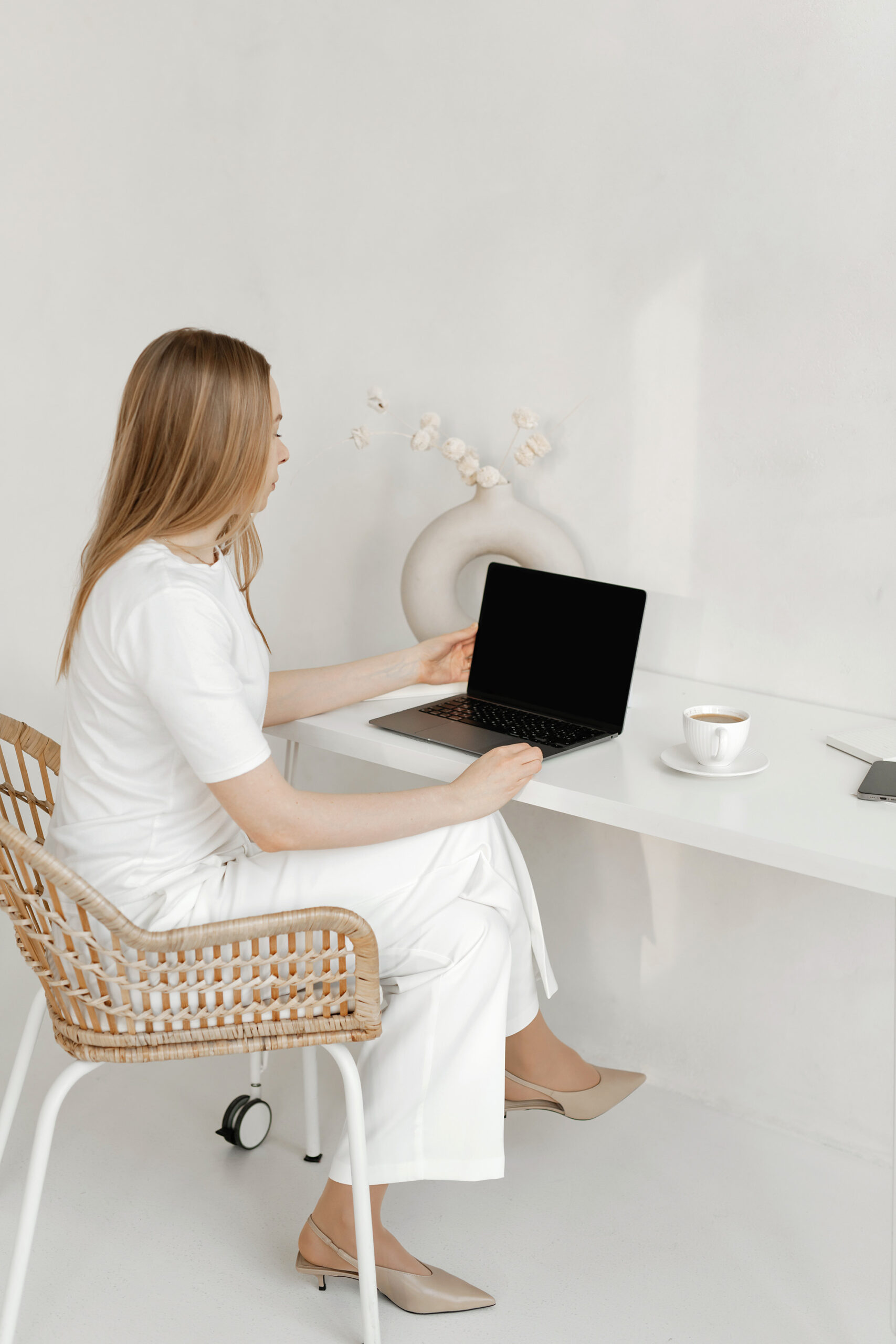 A woman in white clothing sits on a wicker chair at a white desk, using her laptop to browse a website platform for wedding planners. A cup on a saucer and a vase with dried flowers decorate the serene workspace.